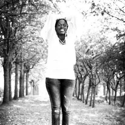 Black-and-white photograph of Tye smiling with her arms raised along a tree-lined path